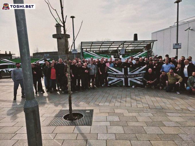 A group of people gathered outdoors holding a Union Jack banner that reads 'UNITED KINGDOM, NEVER IGNORED', with a 'TOSHI.BET' watermark in the corner.