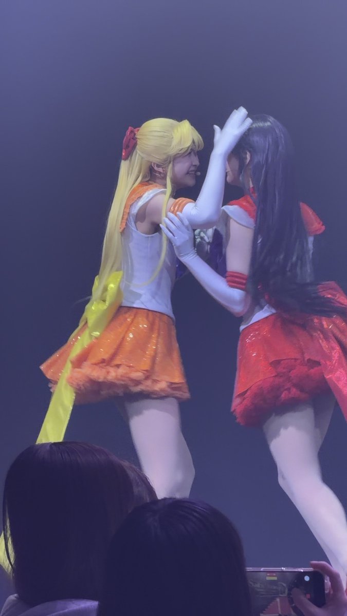 Two performers in Sailor Moon-style costumes—one in an orange skirt and blonde pigtails, the other in a red skirt with long dark hair—interact on stage while an audience member records with a phone.