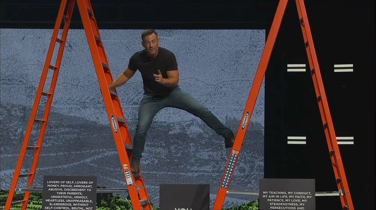 A man in a black shirt and jeans poses dynamically between two orange ladders on a stage with a textured gray backdrop and black placards with white text.
