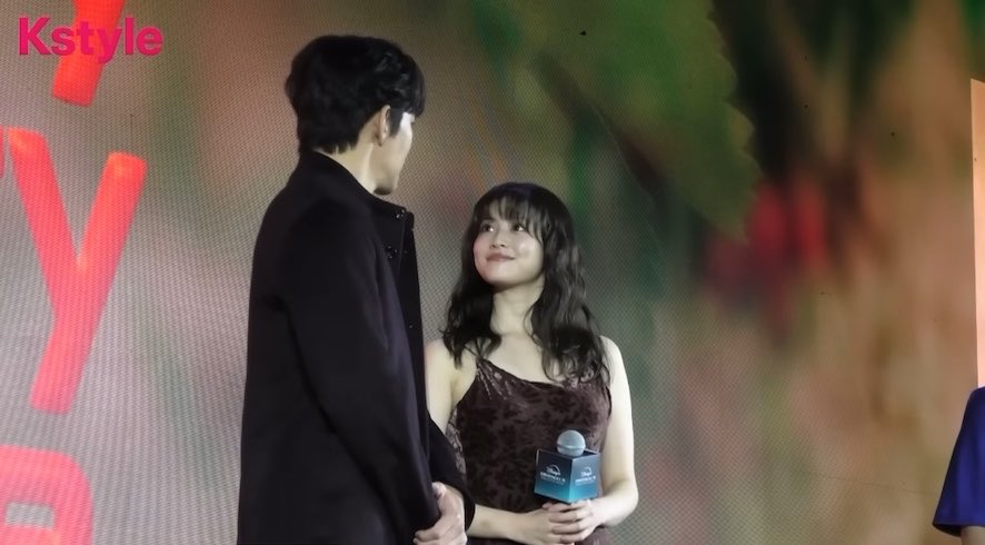 A screenshot from Kstyle showing a man in a black jacket and a smiling woman in a dark velvet dress holding a microphone at an event.