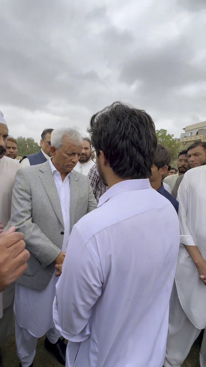 An outdoor gathering showing several men in traditional South Asian attire, with an older man in a checked blazer appearing to be in conversation with a younger man viewed from behind.