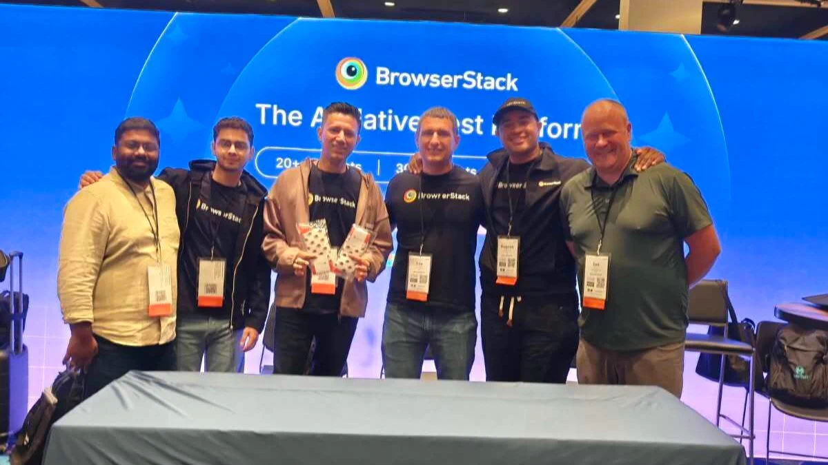 Six men pose smiling at a BrowserStack conference booth, wearing badges; two hold giveaway items in front of a covered table.
