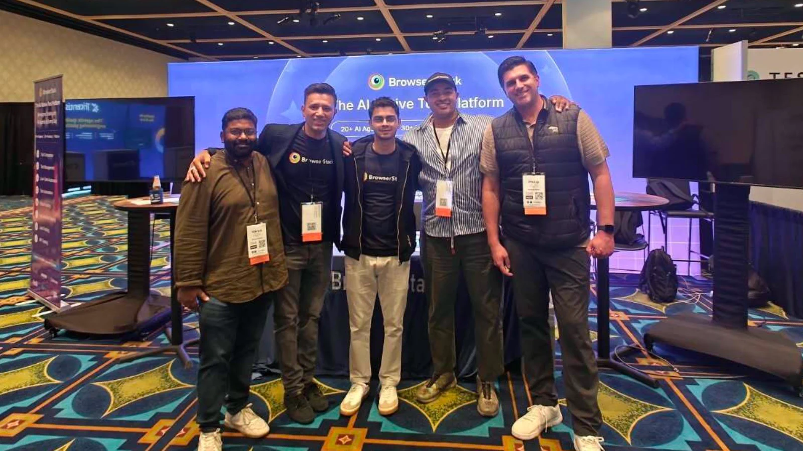 Five men pose arm-in-arm at a tech conference booth for BrowserStack, with large screens and a blue branded backdrop behind them.