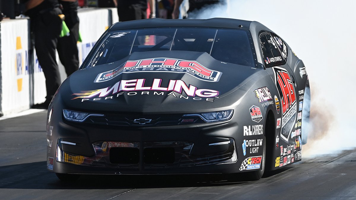 A matte black Chevrolet Camaro Pro Stock race car, adorned with "Melling Performance" branding across the hood and driver's name "Aaron Stanfield" on the side window, performs a smoky burnout at the starting line with numerous sponsor decals including R+L Carriers and Outlaw Light visible on the bodywork.