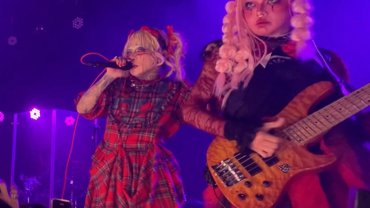 A live concert performance featuring a blonde singer in a red plaid dress with face paint singing into a microphone, alongside a bassist with long white hair playing a wooden bass guitar on a blue-lit stage.