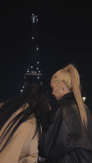 A nighttime photograph of two women standing close together, one with blonde hair in a high ponytail wearing a leather jacket and another with long dark hair, with the Eiffel Tower illuminated in the background.