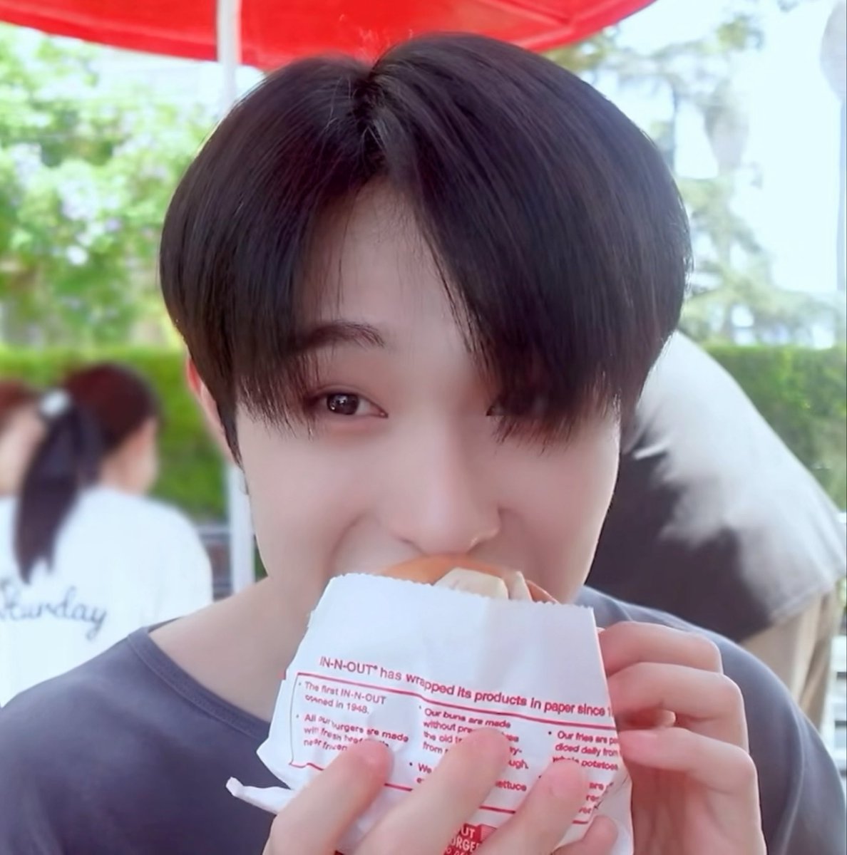 A young person with dark hair eating an In-N-Out burger wrapped in branded paper at an outdoor table under a red umbrella.