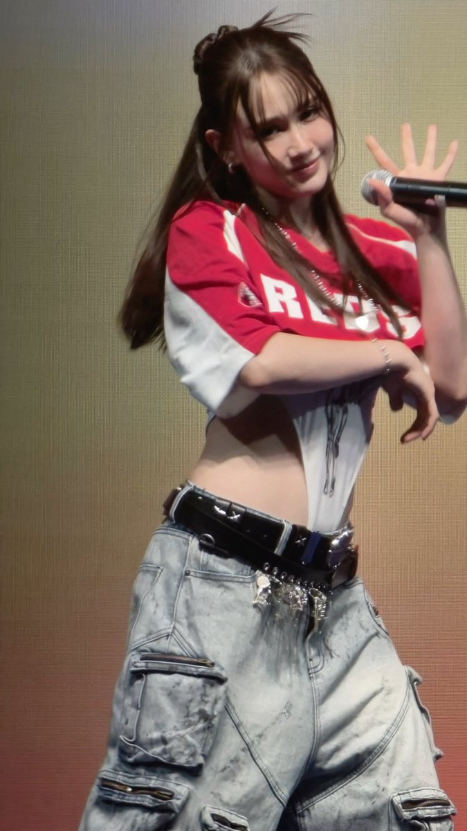 A woman performs on stage wearing a red 'REC' crop top, distressed denim cargo pants with decorative belt, holding a microphone.
