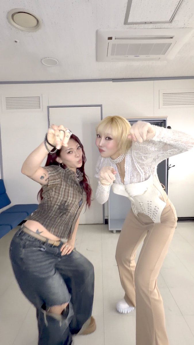 Two women pose playfully for the camera, one with red hair in a plaid top and jeans, and the other with blonde hair in a white lace top and beige pants.