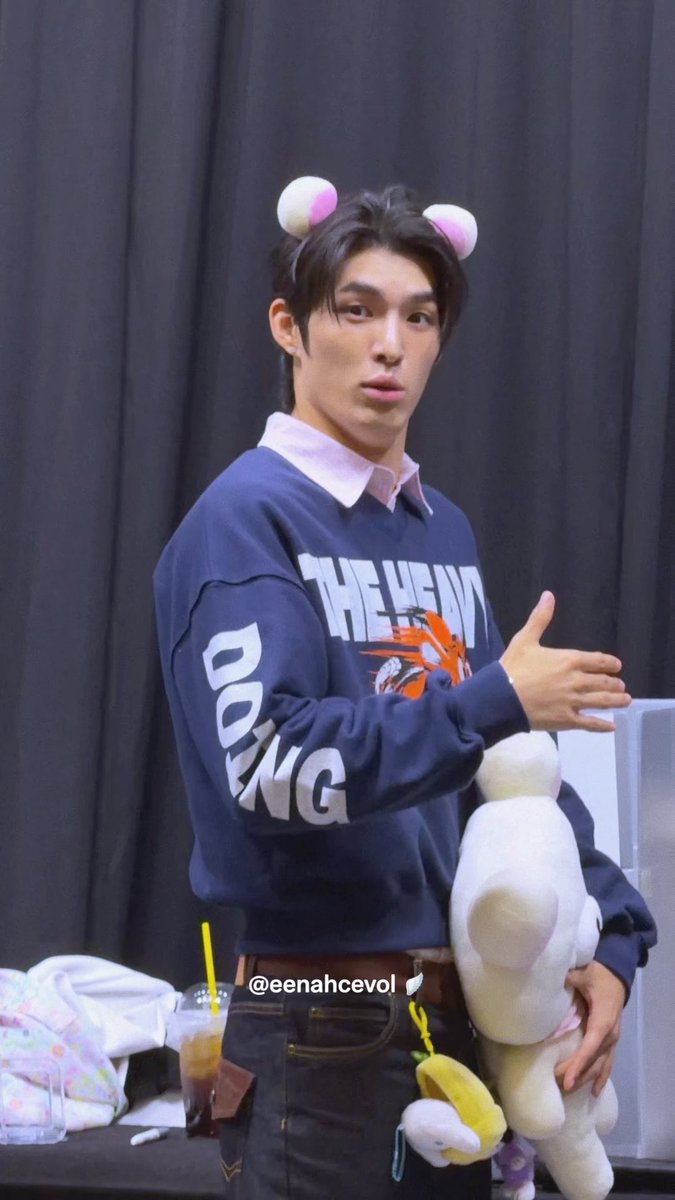 A young man wearing a navy 'THE HEAVY' sweatshirt and cute round ear headband holds a white teddy bear, with @eenahcevol watermark visible.