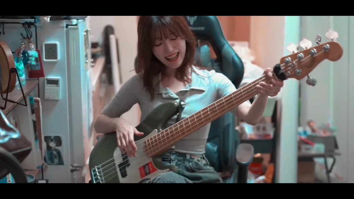 A cheerful young woman with shoulder-length brown hair smiles widely while playing a green bass guitar in a cluttered room filled with music gear and decorations. Her exaggeratedly open mouth and joyful expression playfully match the teasing caption about slurring one's jaw, suggesting she's enthusiastically singing or humming along as she plays.