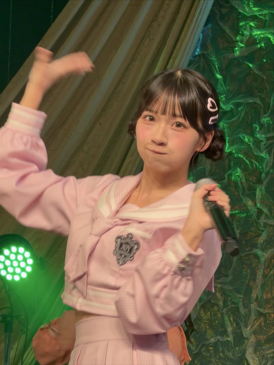 A young Japanese idol performer in a pink sailor-style school uniform holds a microphone on stage, posing with one arm raised against a backdrop of green fabric and stage lighting.