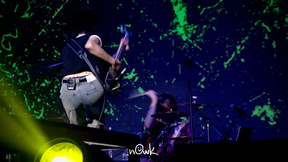 A guitarist stands on stage mid-performance while a drummer plays energetically behind, all illuminated by a vibrant green and blue cosmic backdrop. The watermark "n@wk" appears in the lower center, with yellow stage lighting framing the scene—capturing the passionate "WOOWANZ" (woozzang) concert moment the post celebrates.