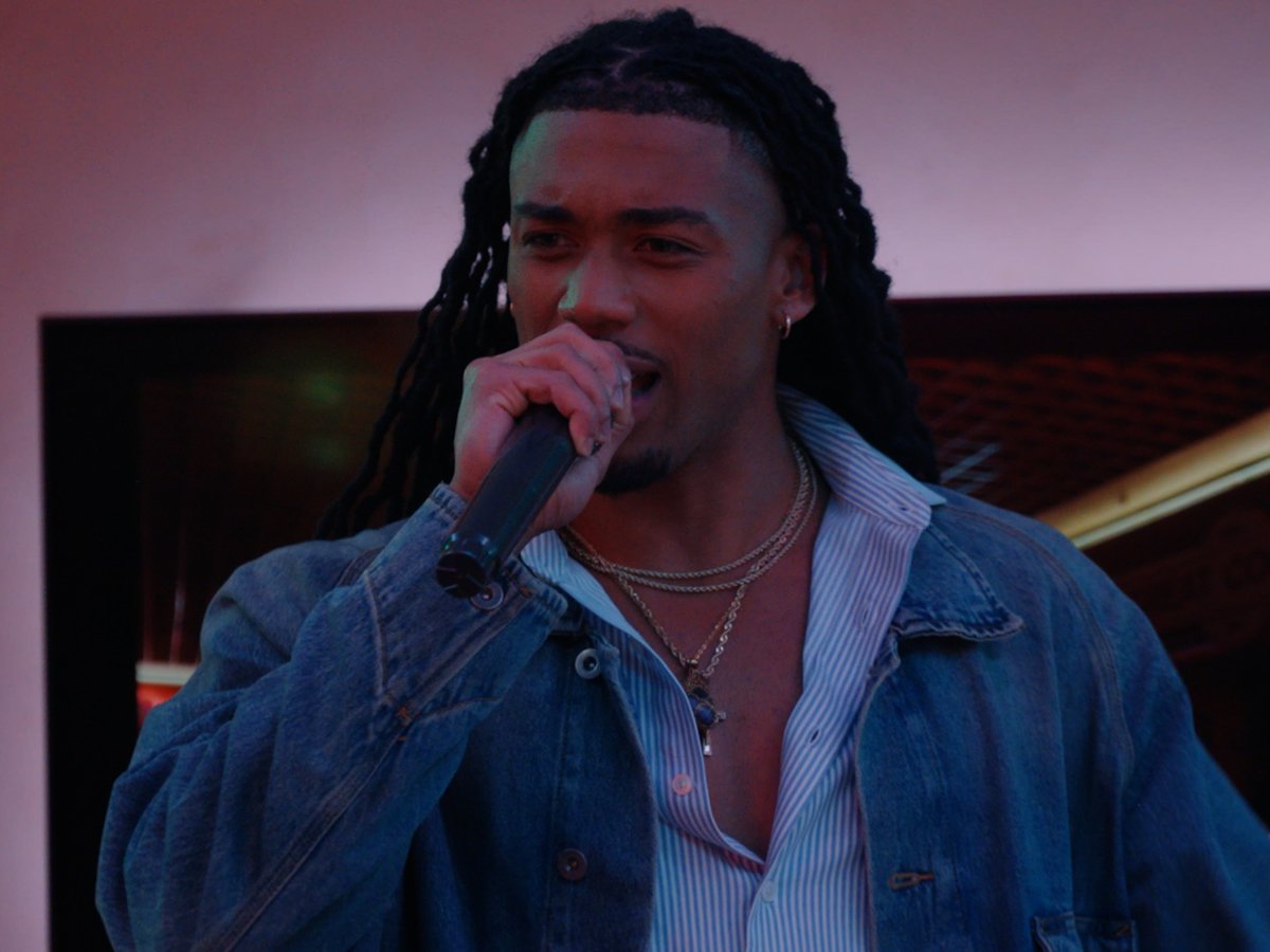 A man with long dreadlocks sings into a microphone while wearing a denim jacket over a striped shirt and layered gold necklaces, set against a dimly lit backdrop with a screen behind him. The caption "just gonna leave this here" suggests a quietly appreciative or teasing post, inviting viewers to simply take in the striking, moody performance moment.