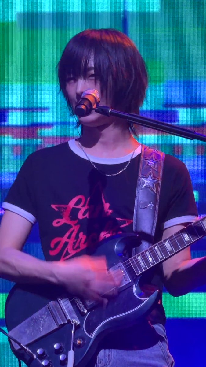 A musician with shoulder-length dark hair performing on stage, singing into a microphone while playing an electric guitar, wearing a black t-shirt with red graphic text and a star-patterned guitar strap.