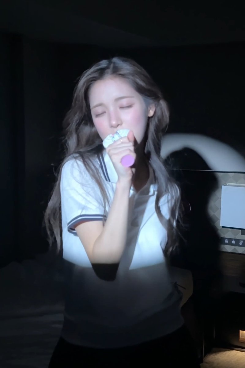 A young woman with long wavy hair sings into a white microphone with a pink base in a dimly lit room, wearing a white polo shirt with dark trim.
