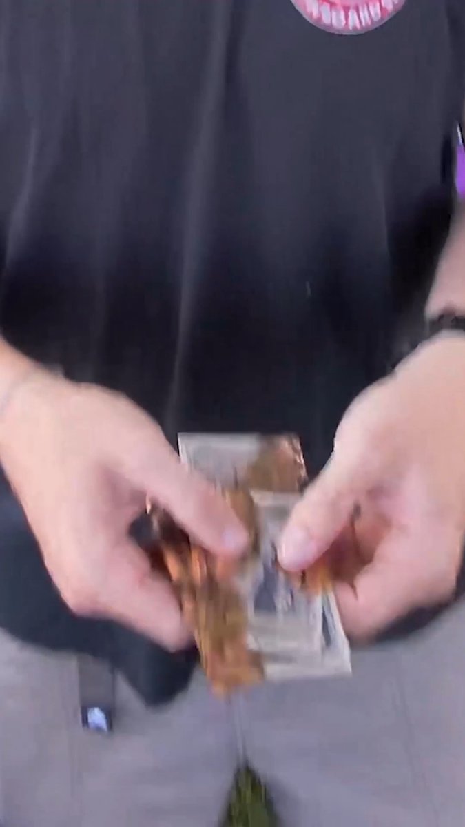 A person wearing a black shirt and gray pants holds a small pack or box of what appears to be trading cards, with a sticker visible on their chest.
