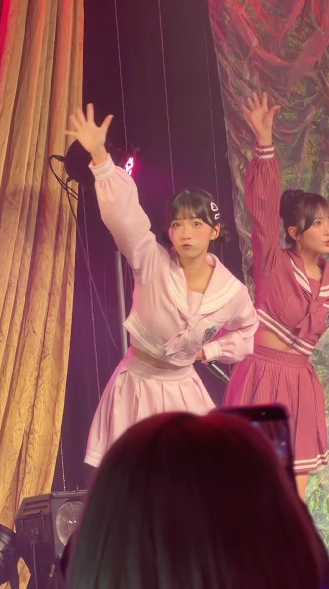 Two young women performing on stage, both with one arm raised; one in a pink outfit and the other in a pink sailor-style uniform.