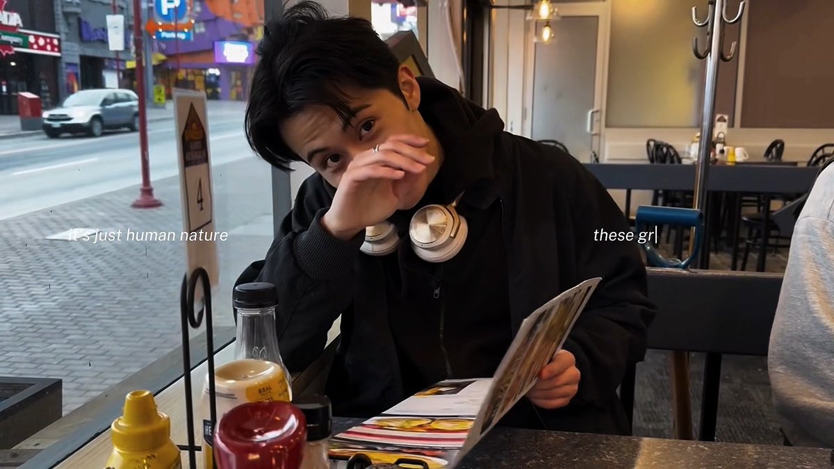 A young man in a black hoodie sits at a restaurant table looking at a menu, with text overlays reading 'It's just human nature' and 'these grl'.