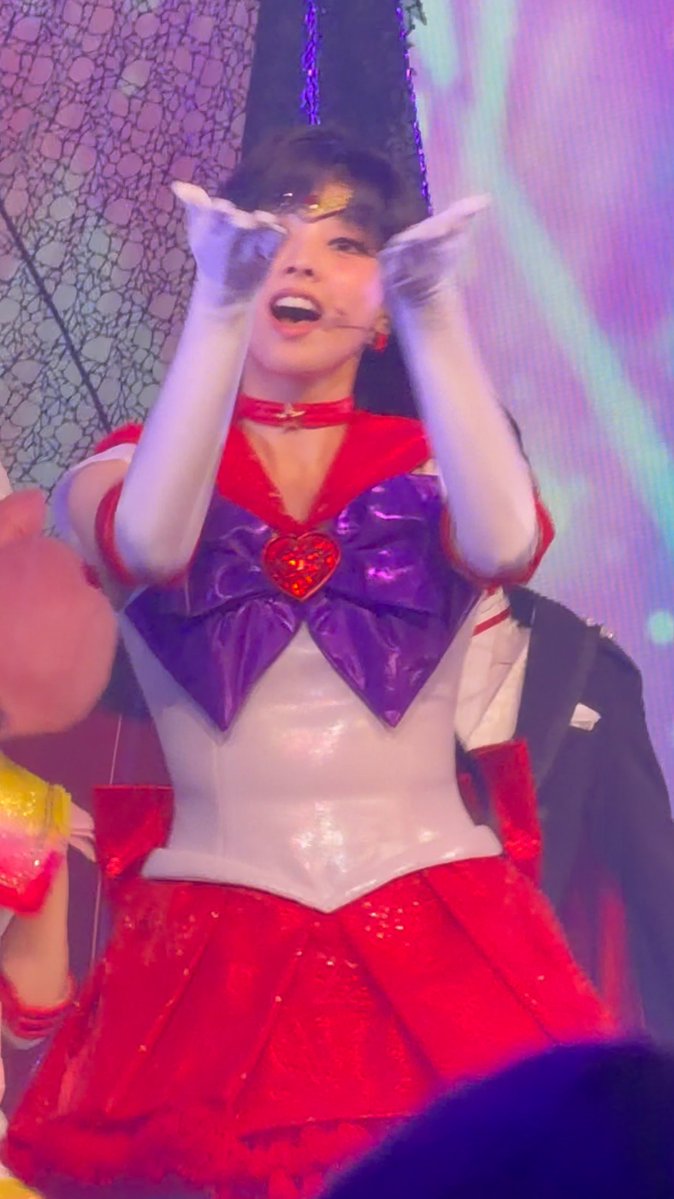 A performer in a Sailor Moon-inspired costume with red skirt, white bodice, and large purple bow poses with gloved hands near their face on a colorful stage.