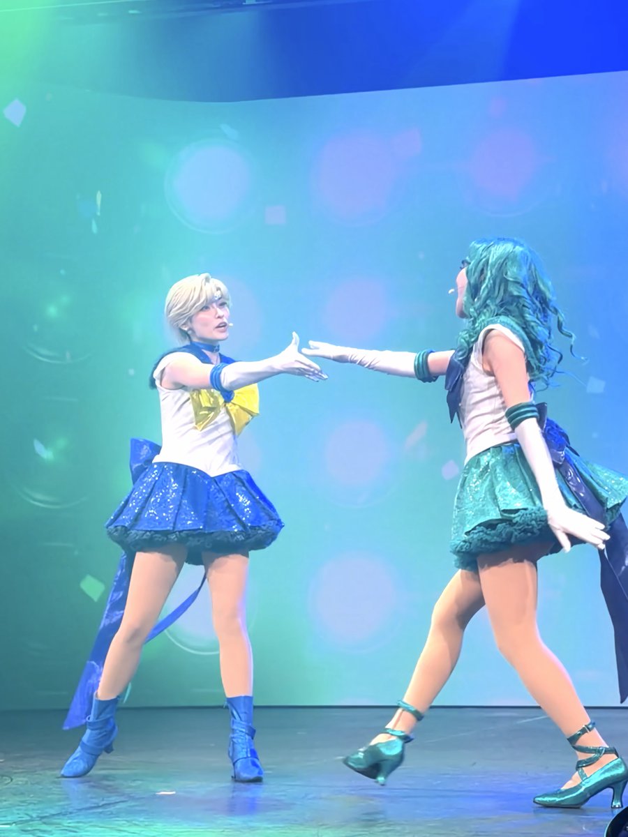 Two performers in Sailor Moon-inspired costumes (one with blonde hair in a blue skirt, one with teal hair in a green skirt) reach toward each other on a stage with blue and green lighting.