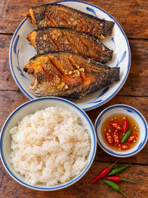 A rustic wooden table holds a blue-rimmed plate of three fried fish, a bowl of white rice, and a small dish of dipping sauce with sliced red and green chilies.