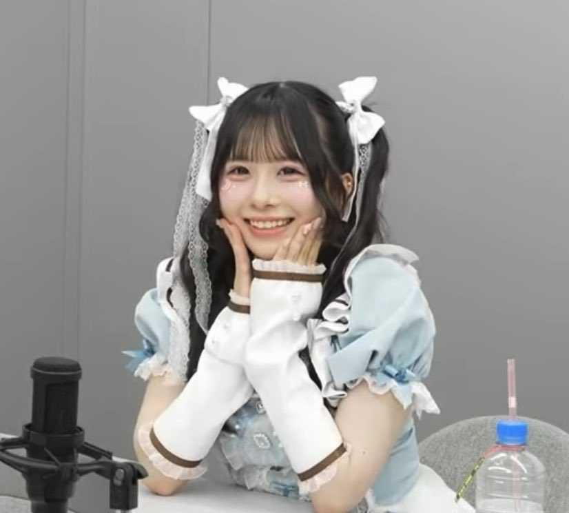A young woman with long dark hair styled in twin tails with white ribbons smiles brightly while resting her chin on her hands, wearing a frilly pastel blue and white outfit resembling a maid costume, with a microphone and water bottle visible nearby.