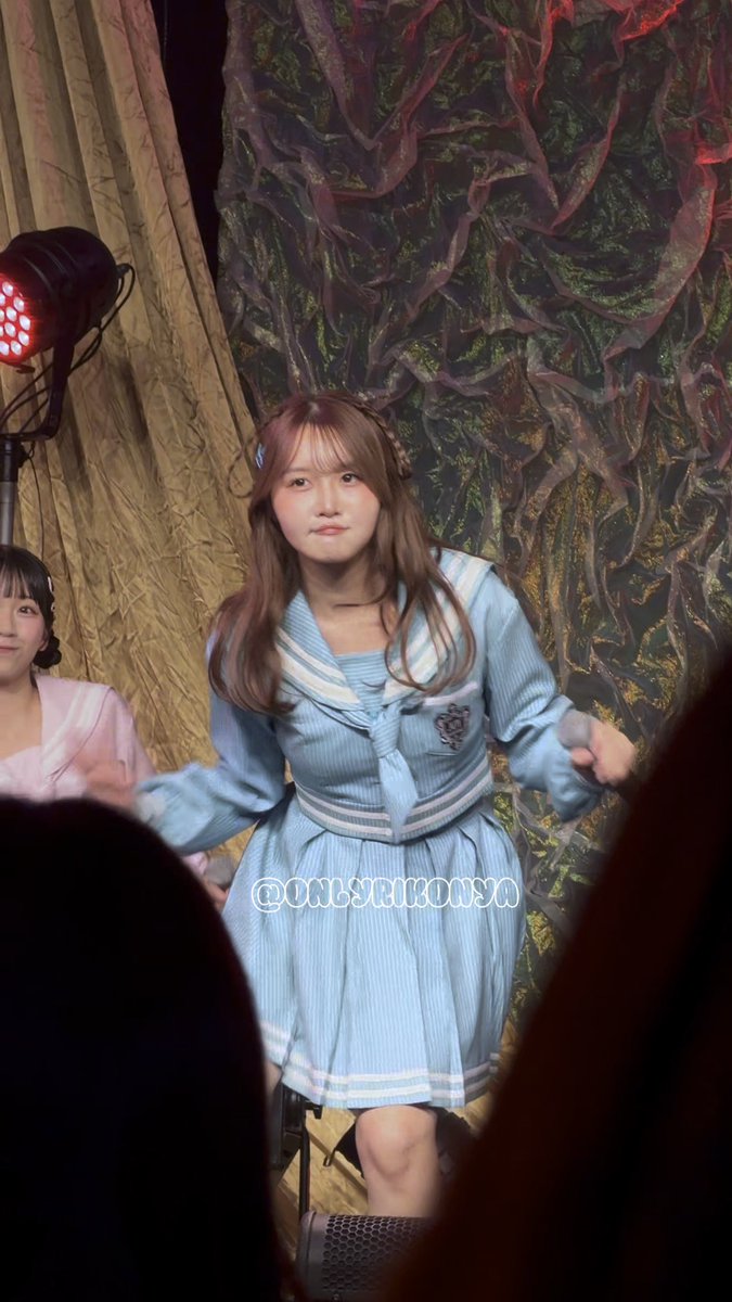 A young woman in a light blue sailor-style school uniform dress performs on stage with an elaborate dark backdrop, with the watermark '@onserikoisa' visible.