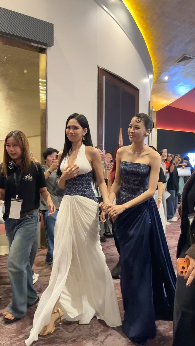 Two elegantly dressed women, one in a white and blue gown and another in a navy strapless dress, hold hands at what appears to be a formal event.