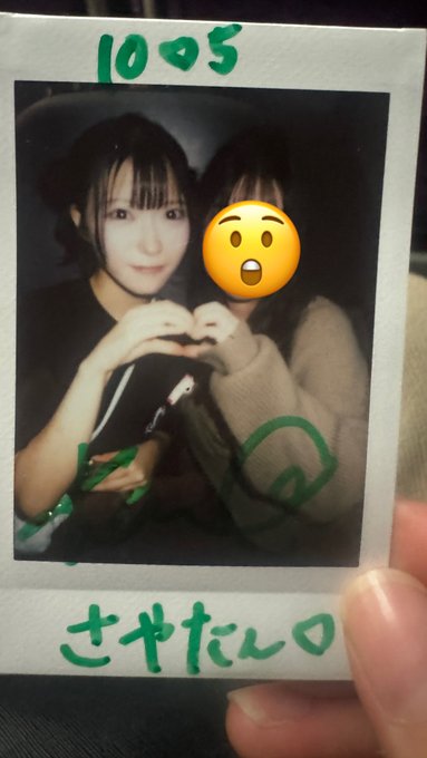 A Polaroid-style photo of two young women forming a heart shape with their hands together, dated "10♡5" with a Japanese signature "さきたん" written in green marker at the bottom, and one face covered by a shocked emoji.