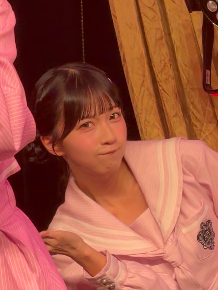 A young woman with dark hair and bangs wearing a pink striped outfit with a decorative emblem, smiling softly indoors.