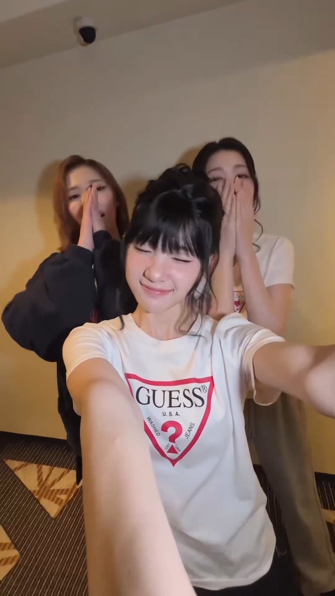 Three women pose for a selfie, with the central figure smiling and the two behind her covering their eyes with their hands.