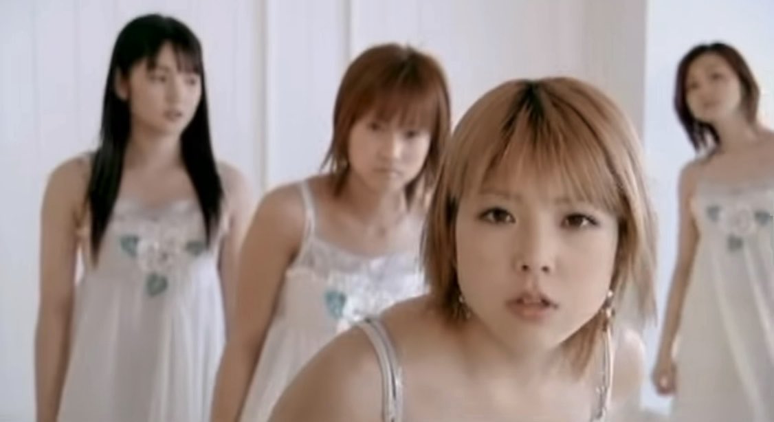 Four young women in matching white dresses with floral embellishments stand in a row, with the front woman in focus and the others blurred in the background.