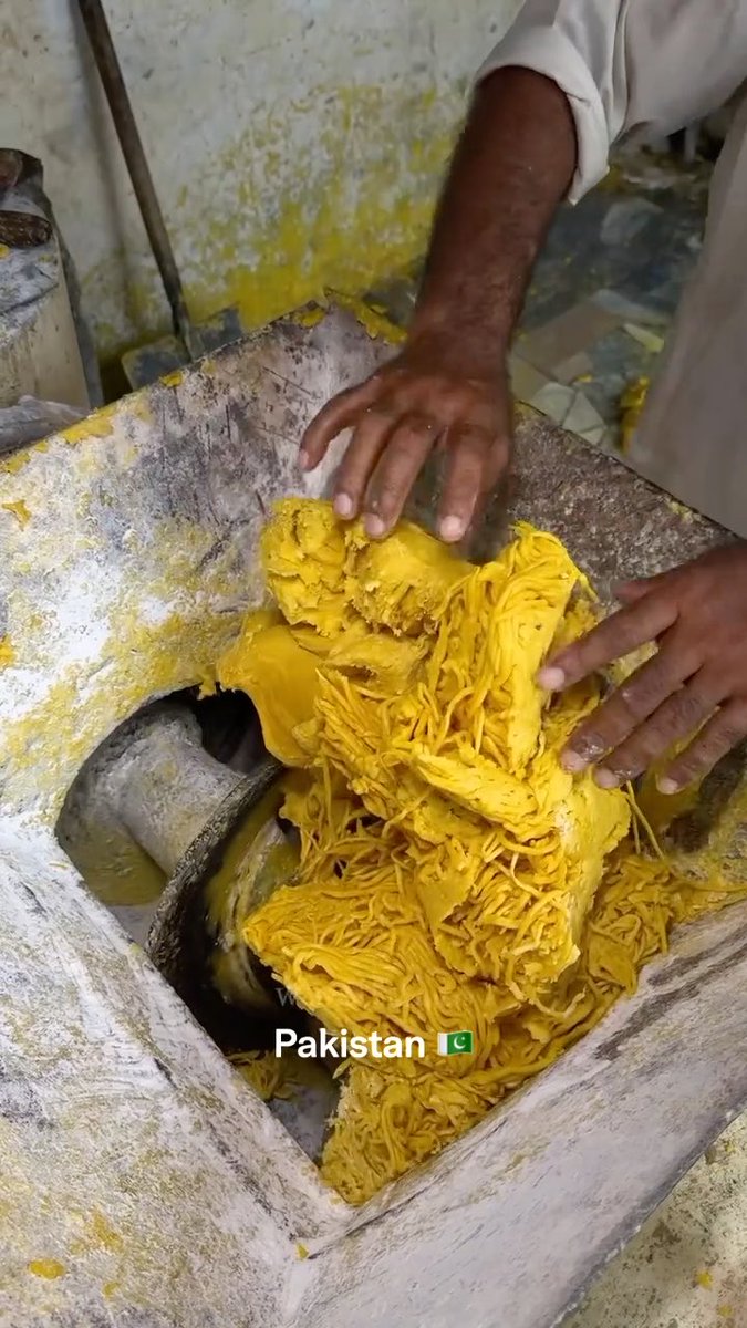 A pair of hands wearing cream-colored sleeves gathers a large mound of bright yellow, stringy food (likely sohan halwa or a similar traditional sweet) from a stone trough, with a "Pakistan 🇵🇰" text overlay visible at the bottom. The image illustrates the post's description of traditional Pakistani food preparation, showcasing the manual, hands-on process of crafting the treat despite the rustic working conditions.