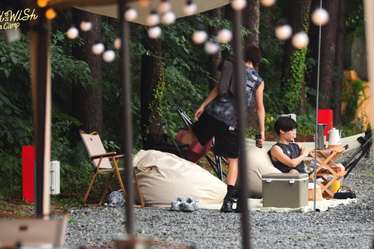 Two young men relax at an outdoor campsite beneath string lights and a canopy, one sitting on a beanbag chair while the other stands nearby.