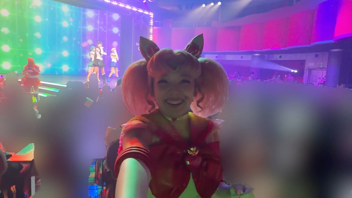 A person in a Sailor Moon cosplay (Chibi Moon) smiling in the foreground, with what appears to be a Sailor Moon performance on stage in the colorful background.
