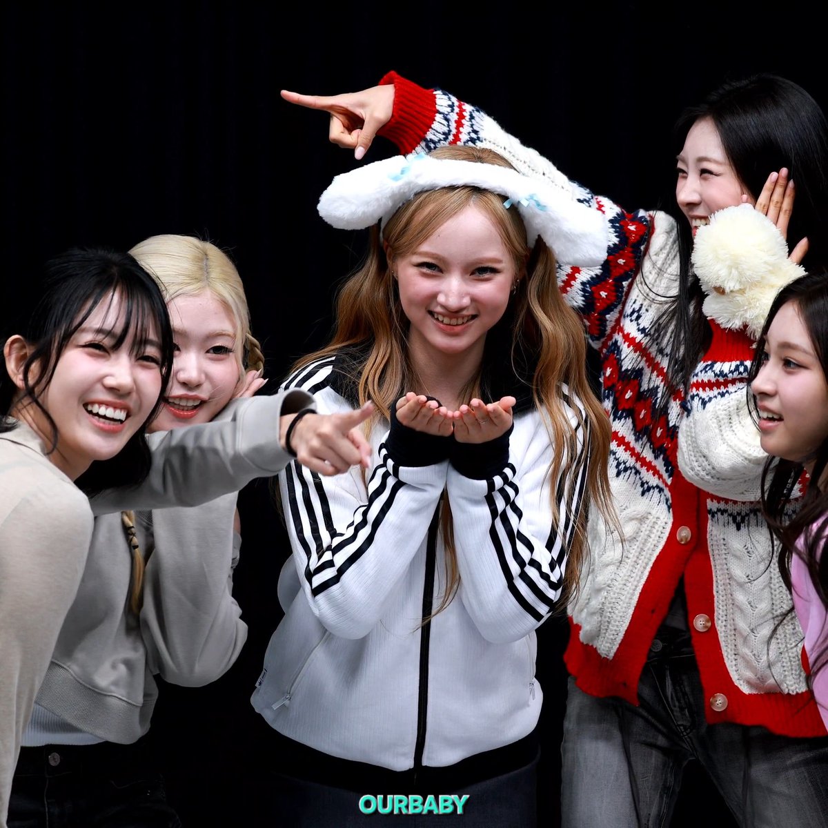 A group photo of five young women smiling and posing, with one wearing a fluffy white bear ear headband and Adidas jacket; the text 'OURBABY' appears at the bottom.