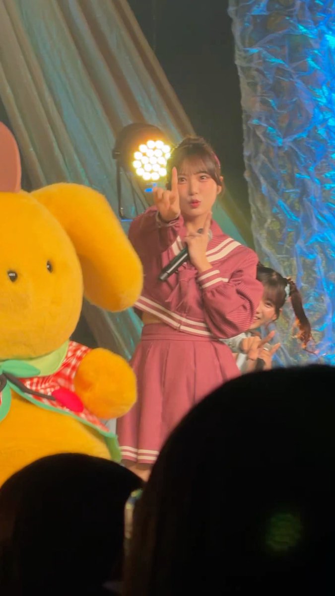 A young woman in a pink outfit performing on stage, holding a microphone and making a peace sign, with a large yellow plush character visible in the foreground.