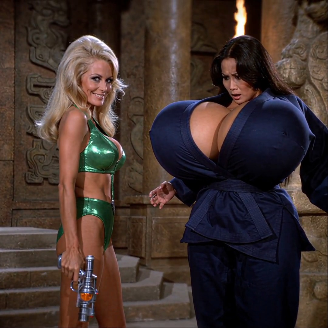 A blonde woman in a sparkly green bikini holds a ray gun while smiling, standing next to a shocked-looking woman wearing an oversized dark blue robe-like outfit with exaggerated proportions, set in an ancient temple setting. The caption "that's a real upgrade!" humorously suggests a dramatic transformation or enhancement, as the woman in the green bikini appears pleased with herself while the other looks dismayed at her new exaggerated figure.