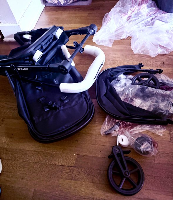Black plastic components of a disassembled "mecool" stroller are scattered on a wooden floor, alongside spare wheels and packaging materials.
