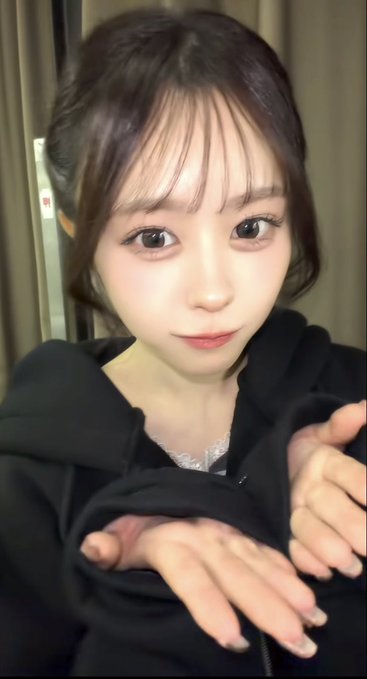 A selfie of a young East Asian woman with dark hair and bangs, wearing a black top, holding her hands out toward the camera.
