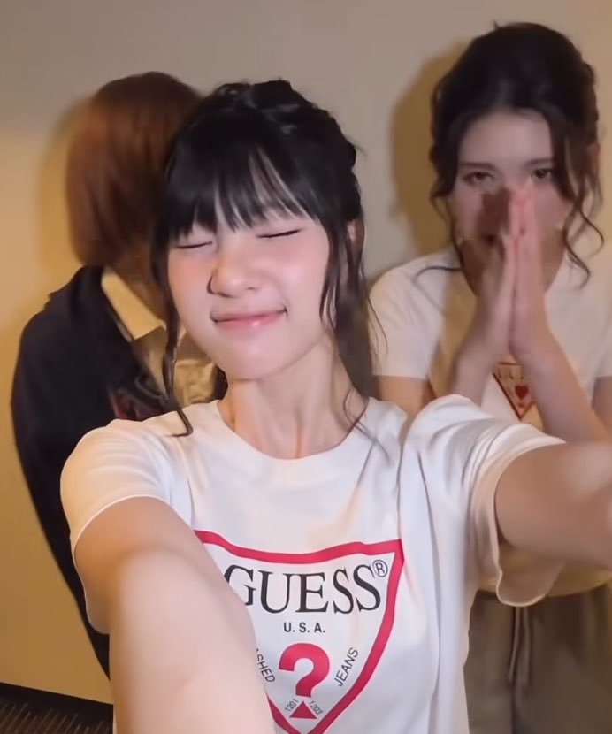 Three young women, likely J-pop idols, with the central figure in a white "GUESS U.S.A." t-shirt smiling with her eyes closed while the one on the right covers her mouth with her hands.