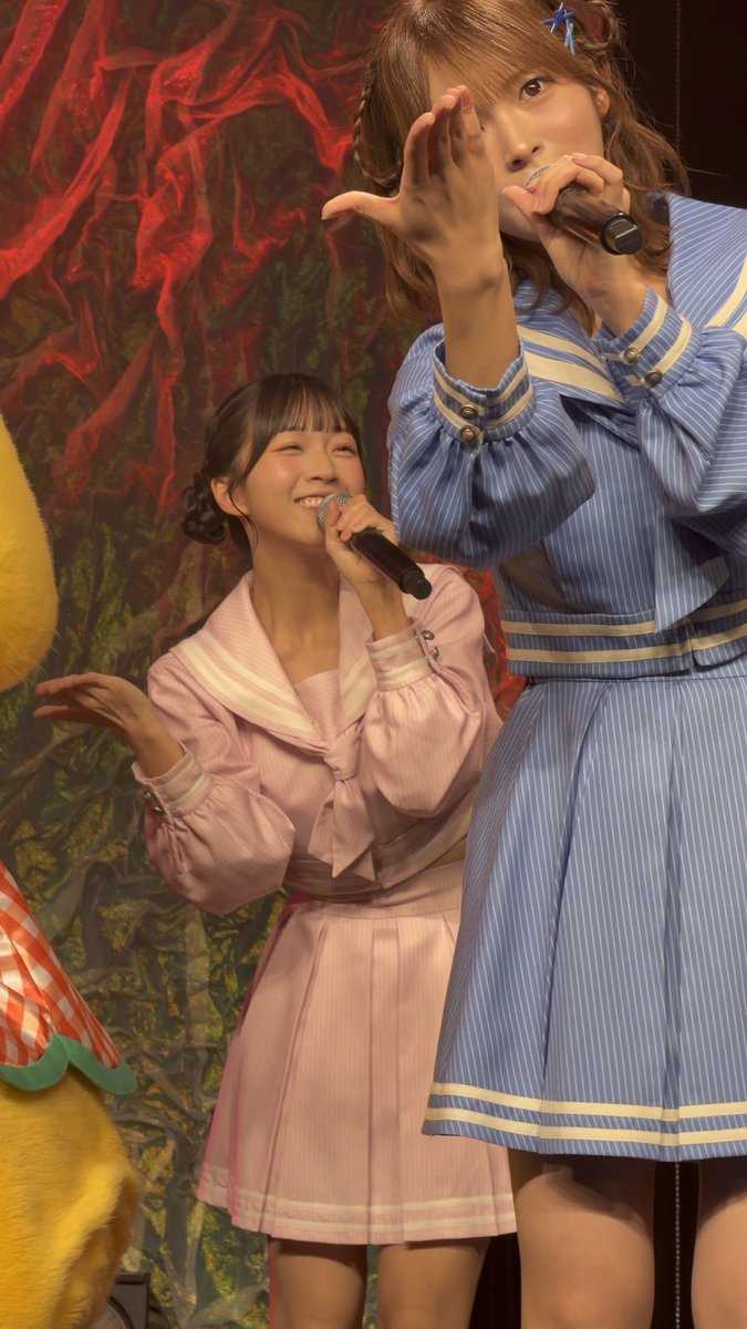 Two young female performers on stage, one in a pink sailor outfit and the other in a blue striped outfit, both holding microphones and smiling during what appears to be a live performance.