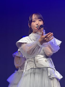 A young woman performing on stage in a white ruffled outfit, singing into a handheld mic while extending her other arm outward against a blue background.