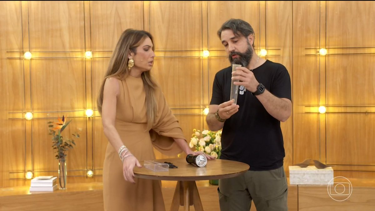 A woman in a tan dress and a bearded man in a black shirt stand behind a small round table examining cylindrical containers during what appears to be a TV show presentation.