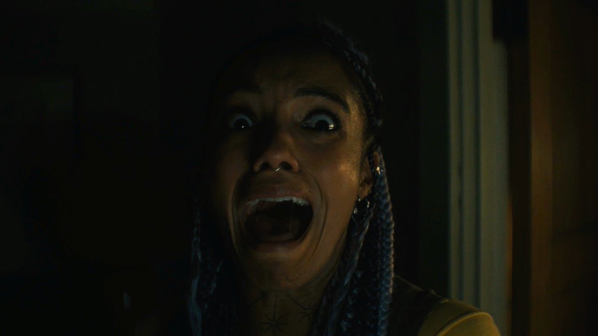 A person with long blue braided hair screams in terror in a dimly lit scene, their wide eyes and open mouth conveying intense fear. This haunting image fits the deleted scene from "Insidious: Out of the Further," capturing a chilling moment of horror as the film prepares for its digital release on October 6 via Sony Pictures at Home.