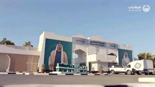 The building prominently displays "SAUDI PUBLIC SECURITY" alongside Arabic script and the royal emblem, featuring large portraits of two Saudi officials in traditional attire, with a white pickup truck parked outside. The image appears to be from a Saudi Public Security social media post honoring teachers for World Teachers' Day, likely set in the security headquarters' courtyard.
