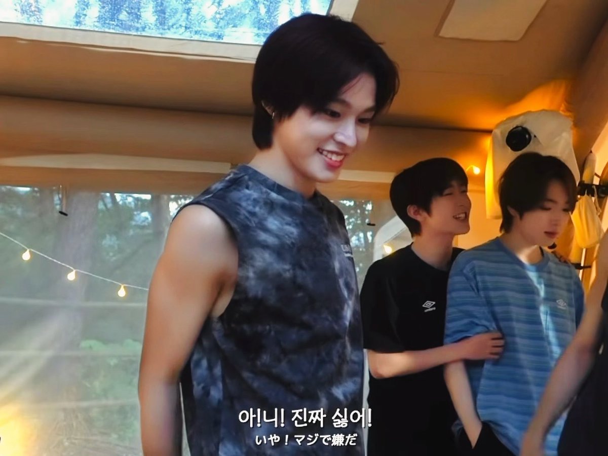 Three young men in casual clothing appear to be in a tent or covered area, with Korean and Japanese subtitles at the bottom of the image.