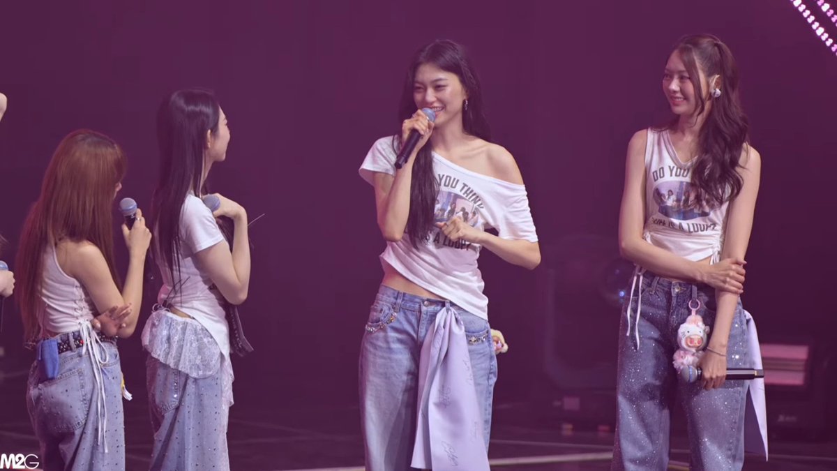 Four women perform on a dimly lit stage with microphones, wearing casual white tops and denim bottoms, with one carrying a small plush charm.