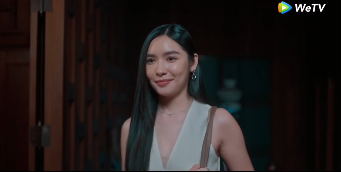 A young woman with long dark hair smiles softly while standing in a dimly lit wooden interior, wearing a white halter top and carrying a brown shoulder bag. The "WeTV" logo appears in the upper right corner, branding the scene as a still from the Thai drama "Khom Klang," fitting the post's reference to Episode 5 of the series.
